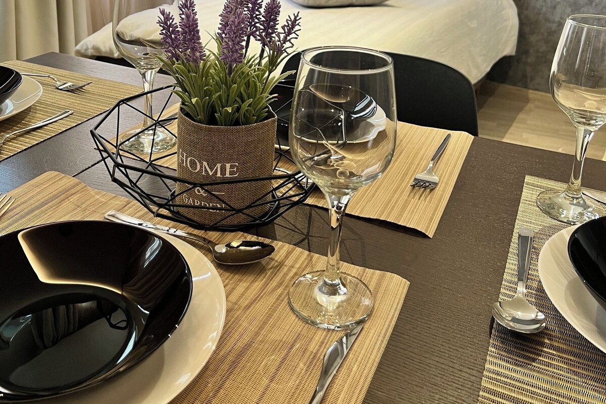 A hotel room with a neatly made bed, a dining table set with plates, glasses, and cutlery, and a small potted plant on the table.