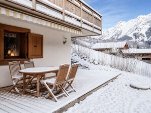 A wooden deck with a table and chairs, a wooden balcony with snow on the railing, and a snowy mountain landscape in the background.