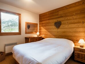 A bedroom with a large bed, wooden panel walls, a window with a view, a bedside table with a lamp, and a wall-mounted heart decoration.