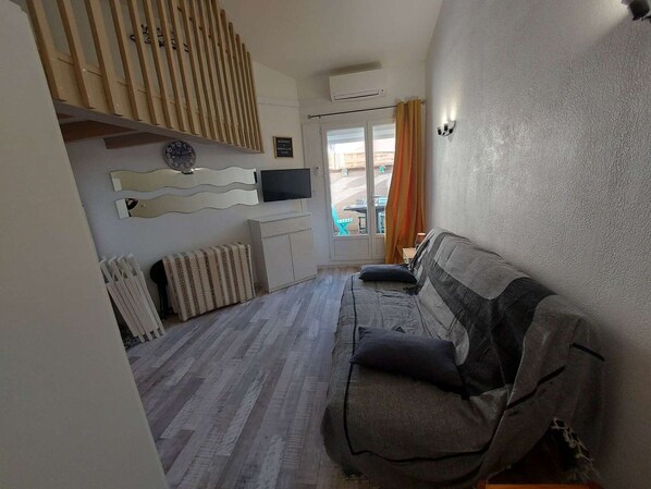 Uma sala de estar com um sofá, uma televisão fixada na parede, teto de ripas de madeira e vista através de uma porta de vidro.
