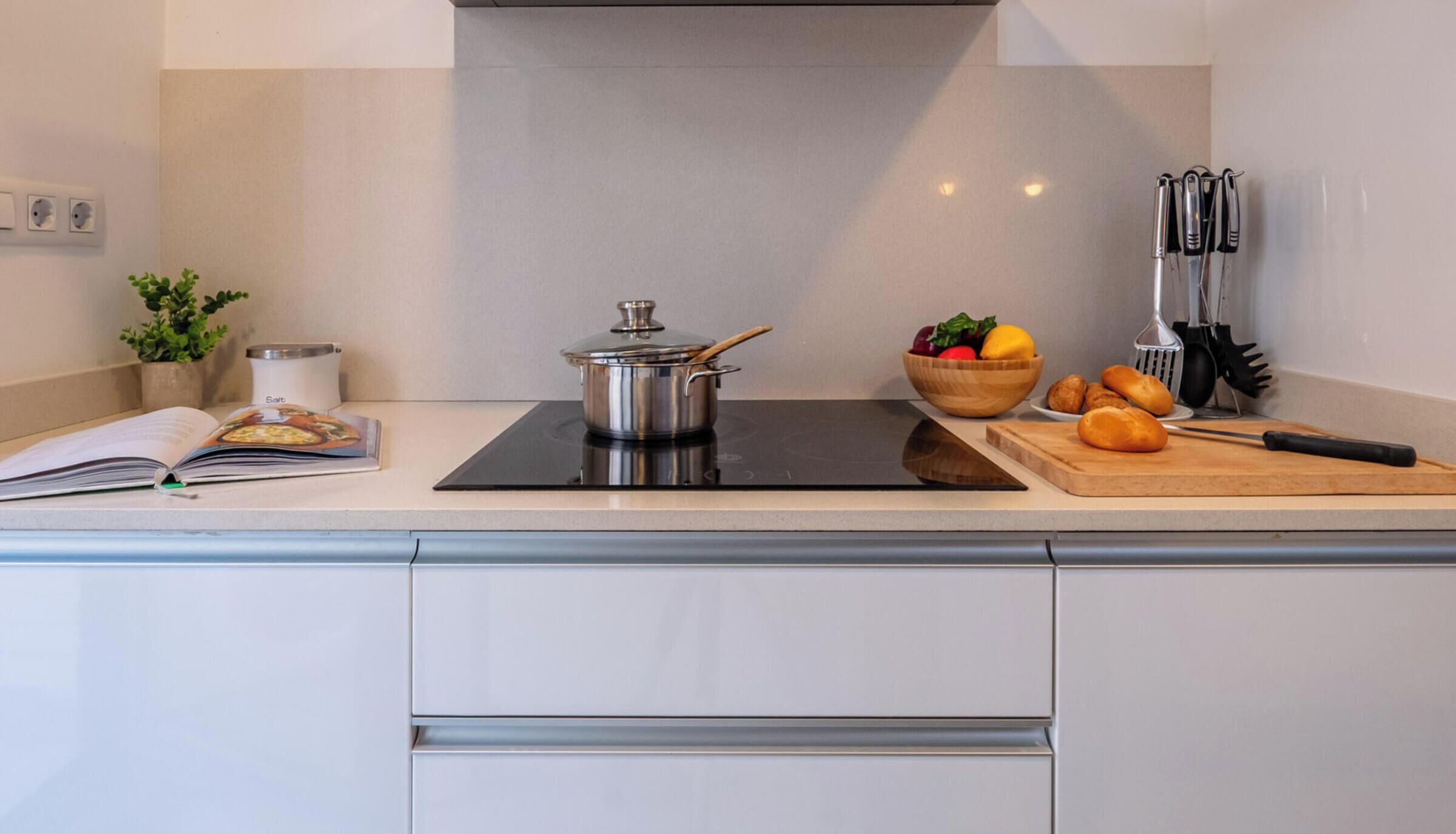 A modern kitchen with a stove, a pot, a bowl of fruit, and a cutting board.