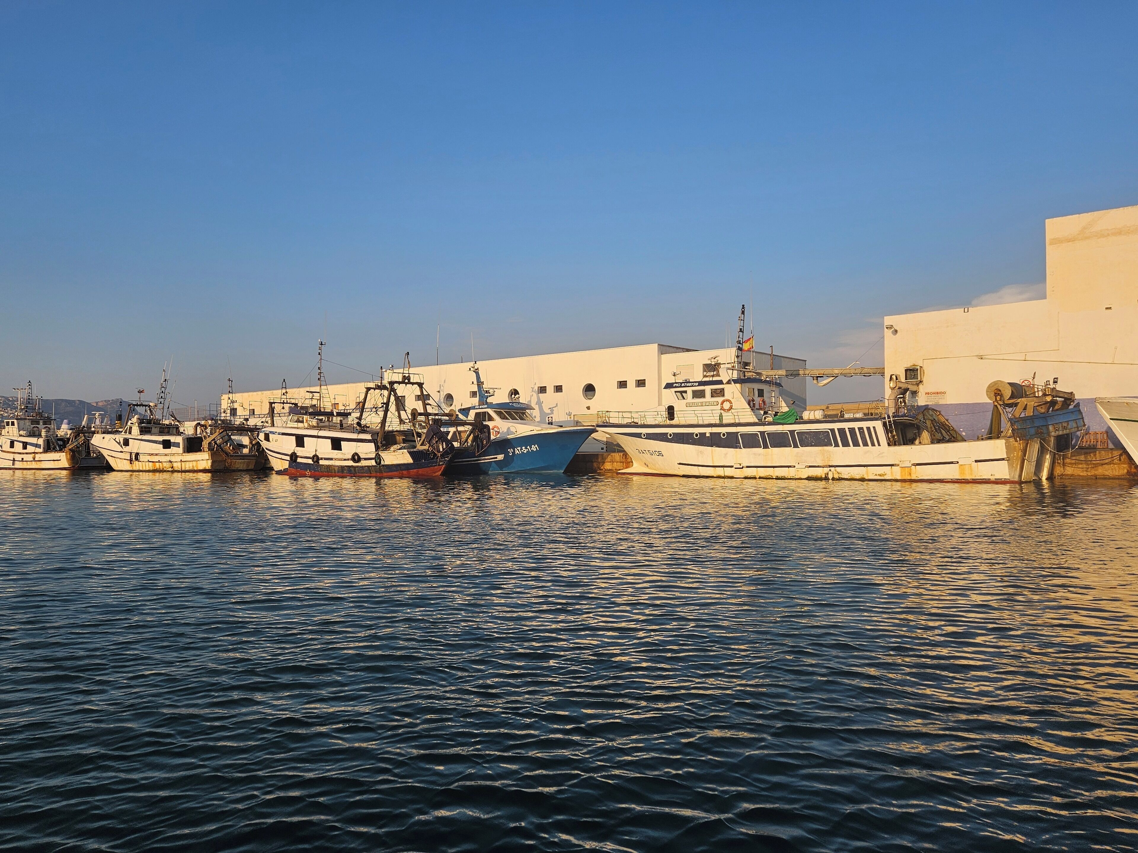 A harbor with several boats docked, a white building with a balcony, and calm water reflecting the boats.