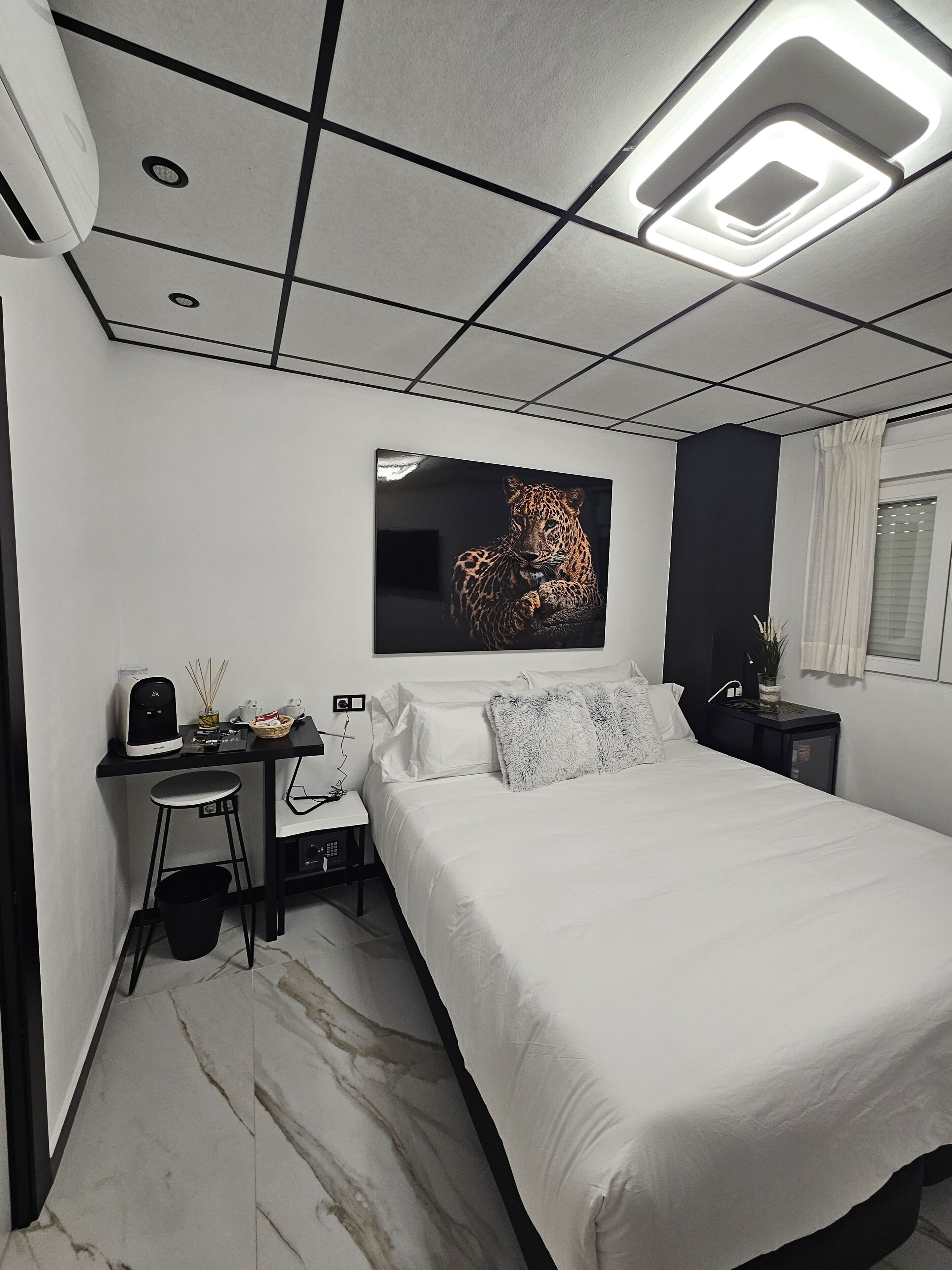 A modern bedroom with a large bed, a desk, and a chair. There is a painting on the wall and a window with curtains.