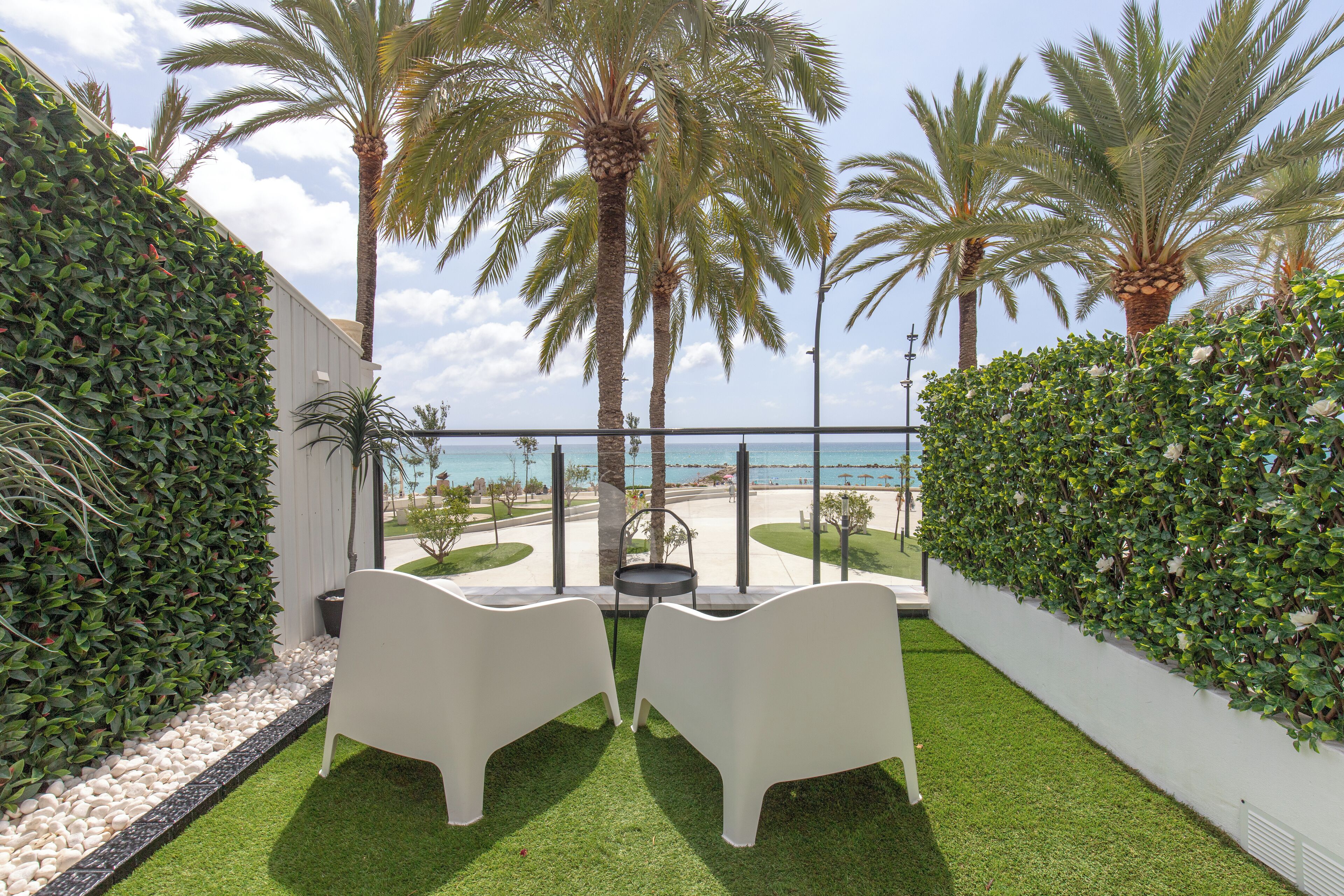 A balcony with two white chairs, a small table, and a view of the beach and palm trees.