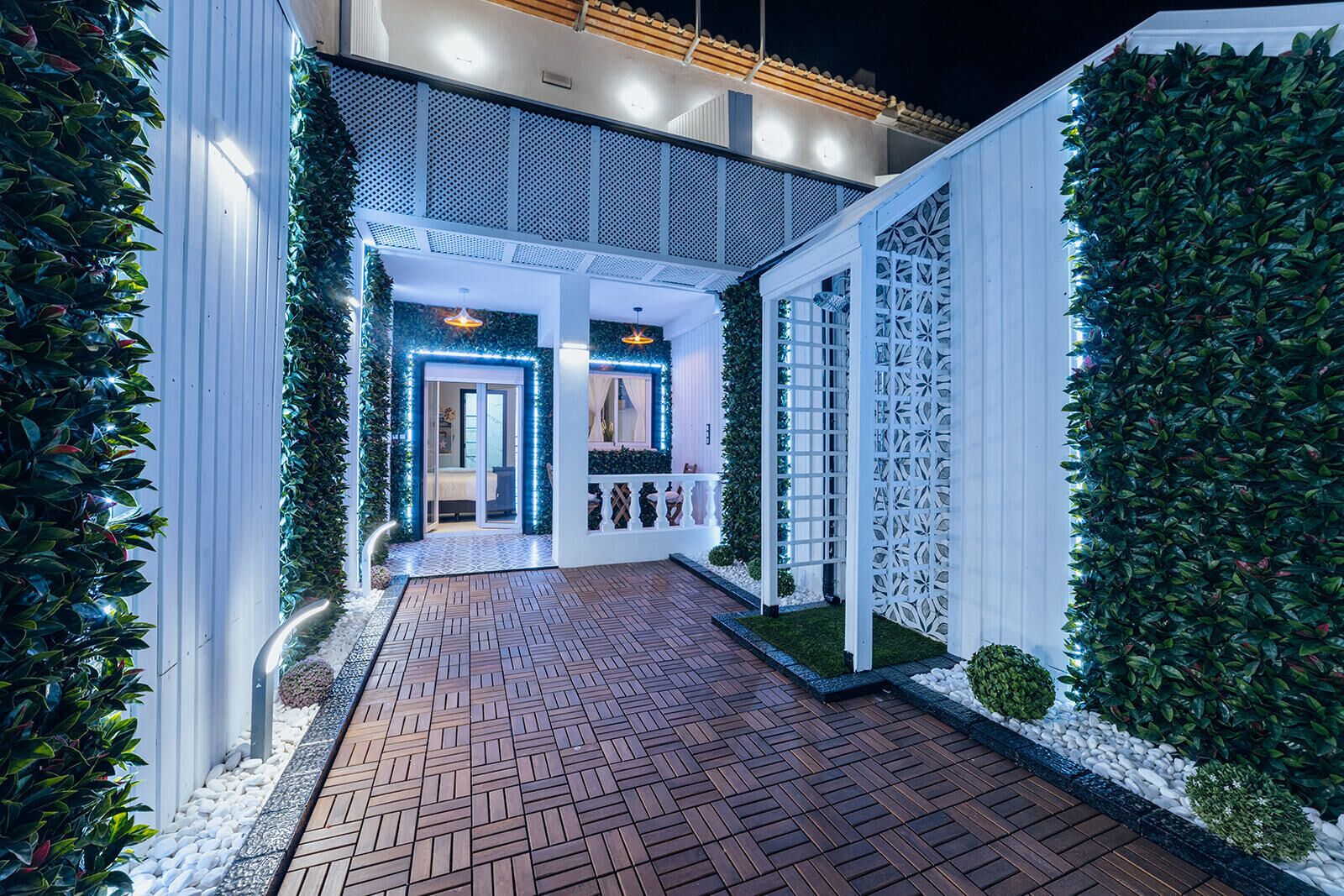 Un edificio moderno con un camino de ladrillo que conduce a una entrada blanca, rodeado de exuberante vegetación y paneles de celosía decorativos.