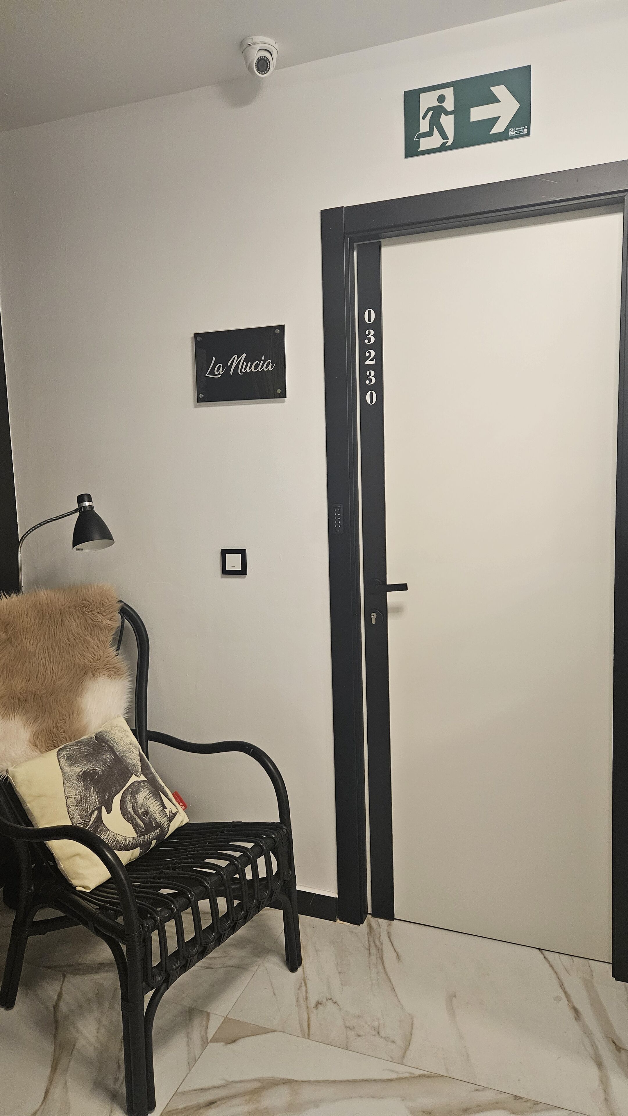 A room with a black metal chair, a fluffy pillow, a wall-mounted lamp, a door with a number, and an emergency exit sign.