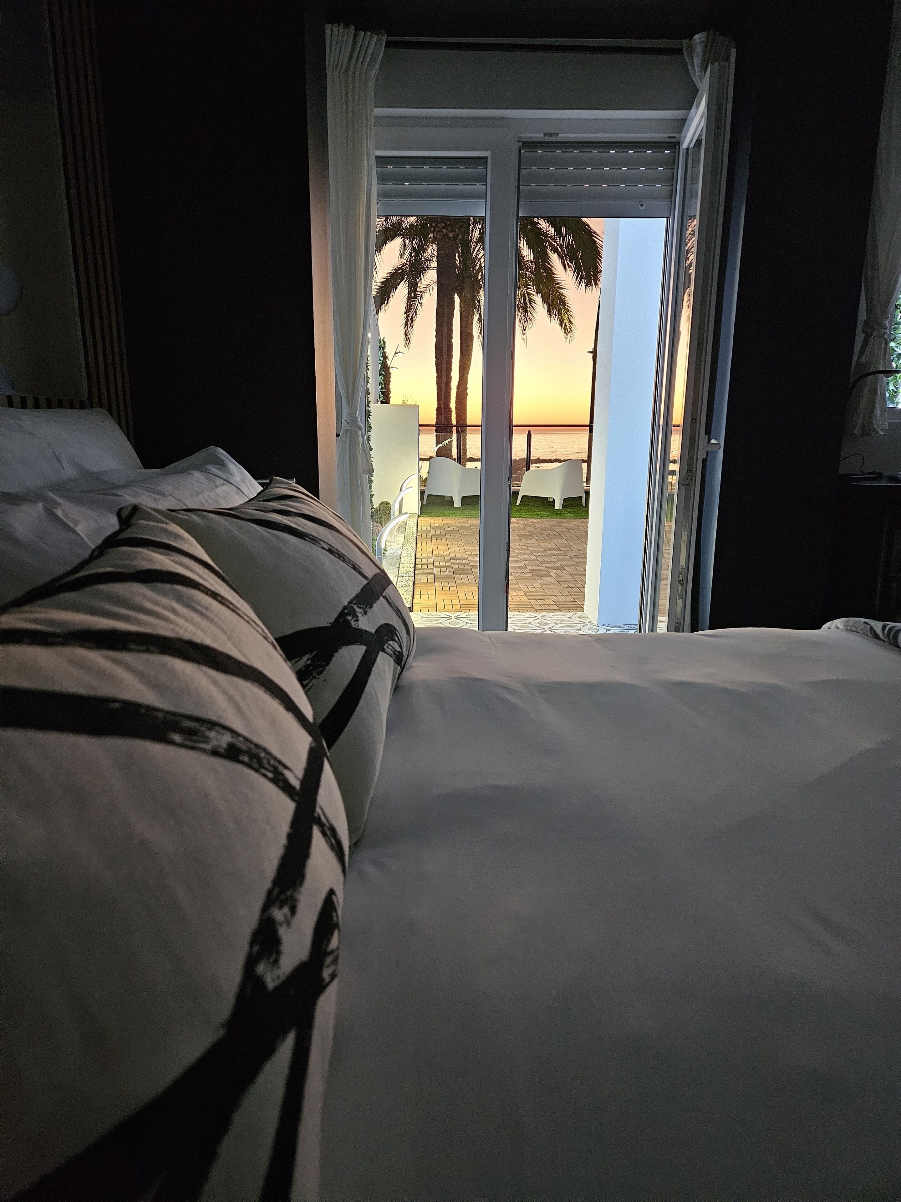 Une chambre avec un lit, une vue sur des palmiers et un coucher de soleil visible à travers une porte ouverte.