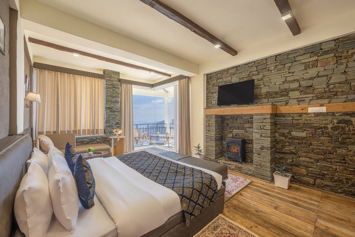 A bedroom with a large bed, a stone fireplace, a TV mounted on the wall, and a balcony with a view.