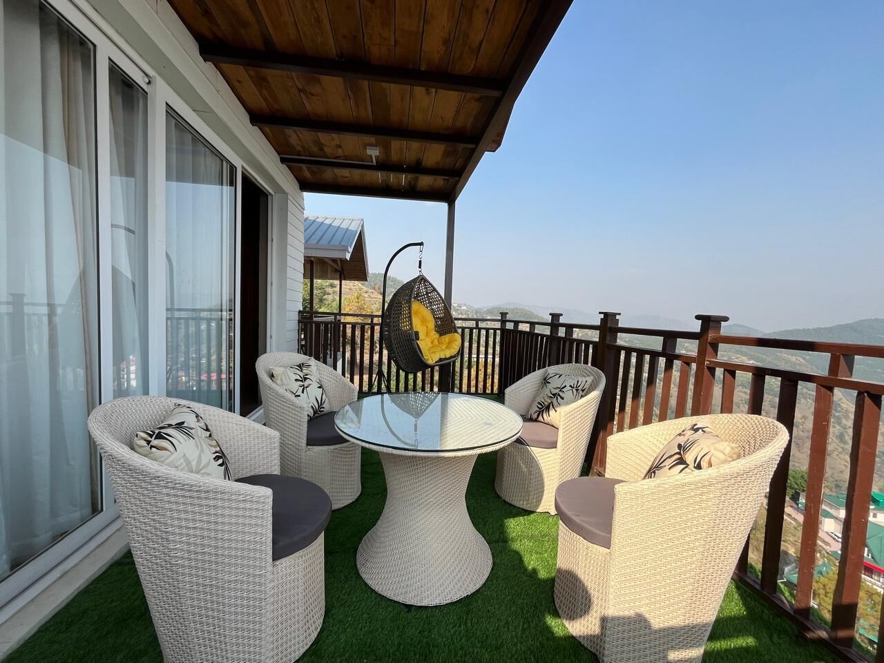 A covered outdoor seating area with wicker furniture and a glass table, overlooking a scenic landscape.
