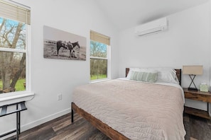 Une chambre à coucher avec un lit, deux fenêtres, un climatiseur fixé au mur, une table de chevet avec une lampe et un tableau représentant des chevaux.