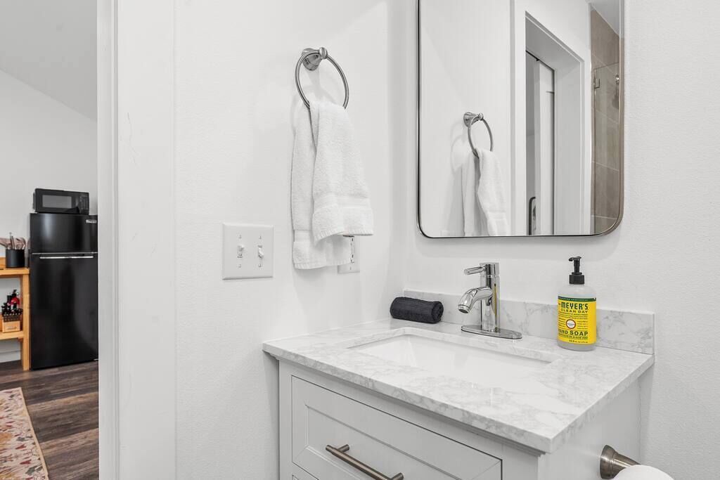 Un baño con un lavamanos blanco, un espejo, un toallero, un dispensador de papel higiénico y una botella de jabón para manos.