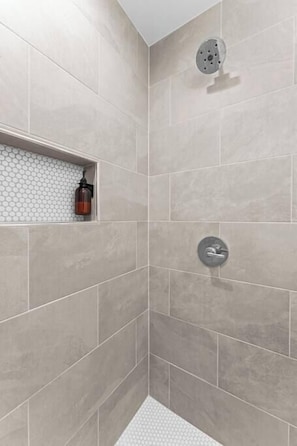 A shower area with beige tiles, a white shelf with a bottle, and a showerhead. - Casita Fulton - Fire Pit & Close to Main St (Fredericksburg)