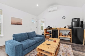 Un salon avec un canapé bleu, une table basse en bois, un coin cuisine avec un réfrigérateur noir et une horloge au mur.