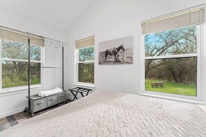 A bedroom with a large bed, a window with a view of trees, a wall-mounted artwork of a horse, and a metal cart with towels. - Casita Fulton - Fire Pit & Close to Main St (Fredericksburg)