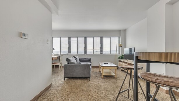 Ein modernes Wohnzimmer mit einem grauen Sofa, einem Holztisch und einem Fernseher. Es gibt ein Fenster mit Jalousien, eine Lampe und eine kleine Pflanze auf dem Fensterbrett.