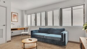 A living room with a blue sofa, a wooden coffee table, a desk with a lamp, and a large window with blinds. - Discover spacious 1 & 2-bedroom apartments & corporate suites in a desirable location (Harrisburg)