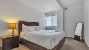 A bedroom with a wooden headboard, a bedside table with a lamp, a window with blinds, and a full-length mirror. - Discover spacious 1 & 2-bedroom apartments & corporate suites in a desirable location (Harrisburg)