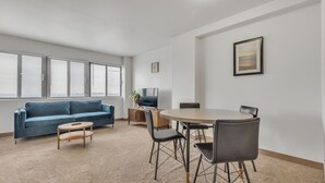 A modern living room with a blue sofa, a small coffee table, a TV on a wooden stand, and a dining table with chairs. - Discover spacious 1 & 2-bedroom apartments & corporate suites in a desirable location (Harrisburg)