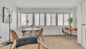Una habitación moderna y luminosa con una mesa redonda, cuatro sillas, un escritorio con computadora y un amplio ventanal con persianas.