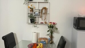 Un comedor con una mesa de vidrio, dos sillas negras, un estante blanco con plantas, un jarrón con flores y un microondas sobre un mueble.
