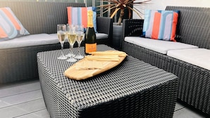 Un divano in vimini con cuscini a righe, una bottiglia di Veuve Clicquot e tre bicchieri di vino bianco su un tavolo.