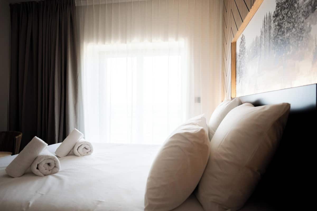 A hotel room with a neatly made bed, rolled towels, and a large window with sheer curtains.