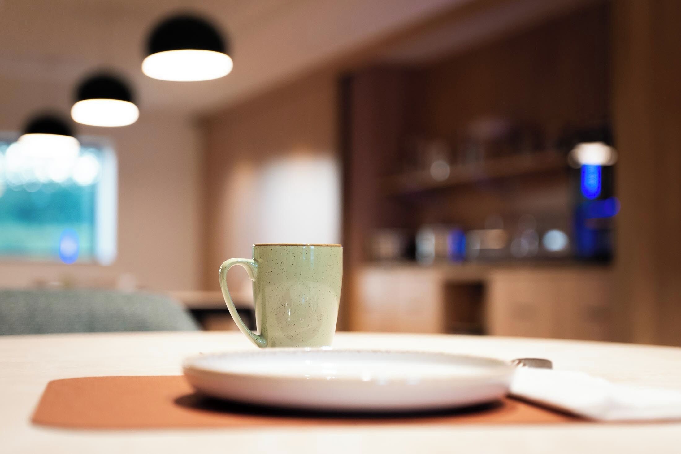 A green ceramic mug on a white plate, a brown placemat, and a white napkin on a wooden table in a room with blurred background featuring shelves and hanging lights.
