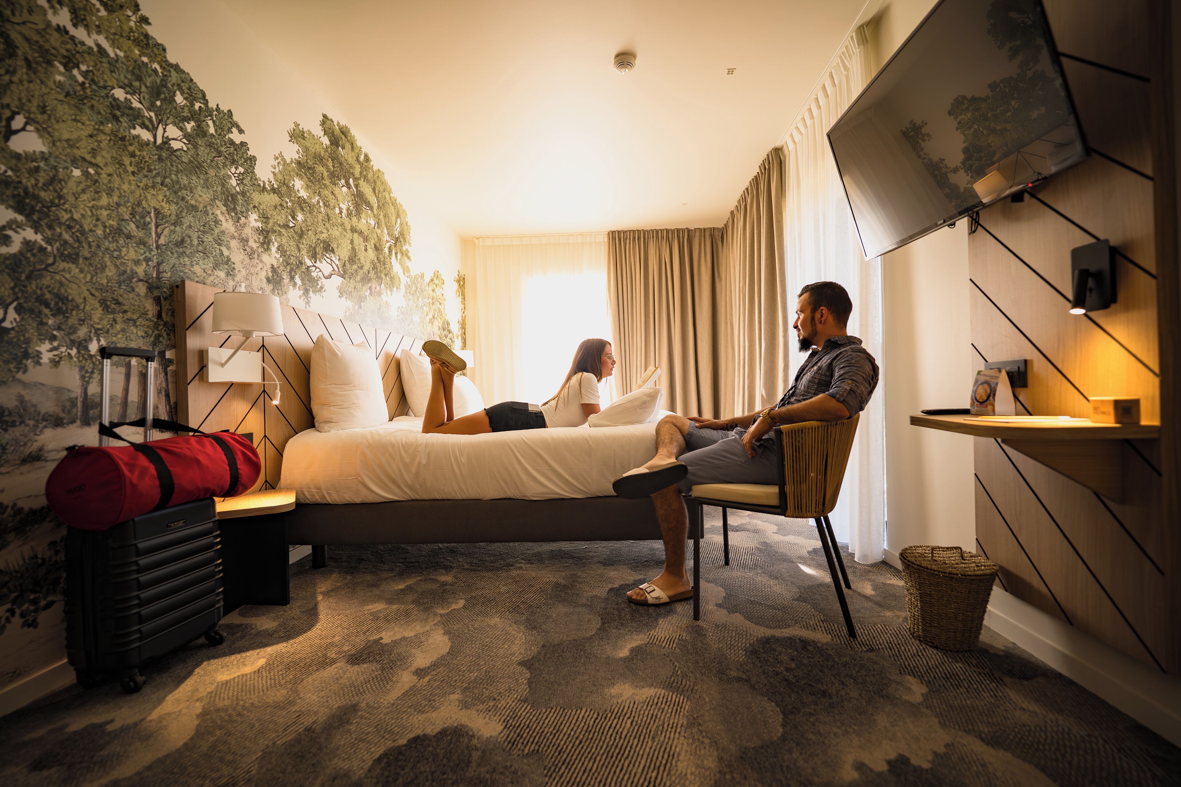 Ein Hotelzimmer mit Bett, Schreibtisch, Stuhl, Koffer und einem an der Wand befestigten Fernseher.
