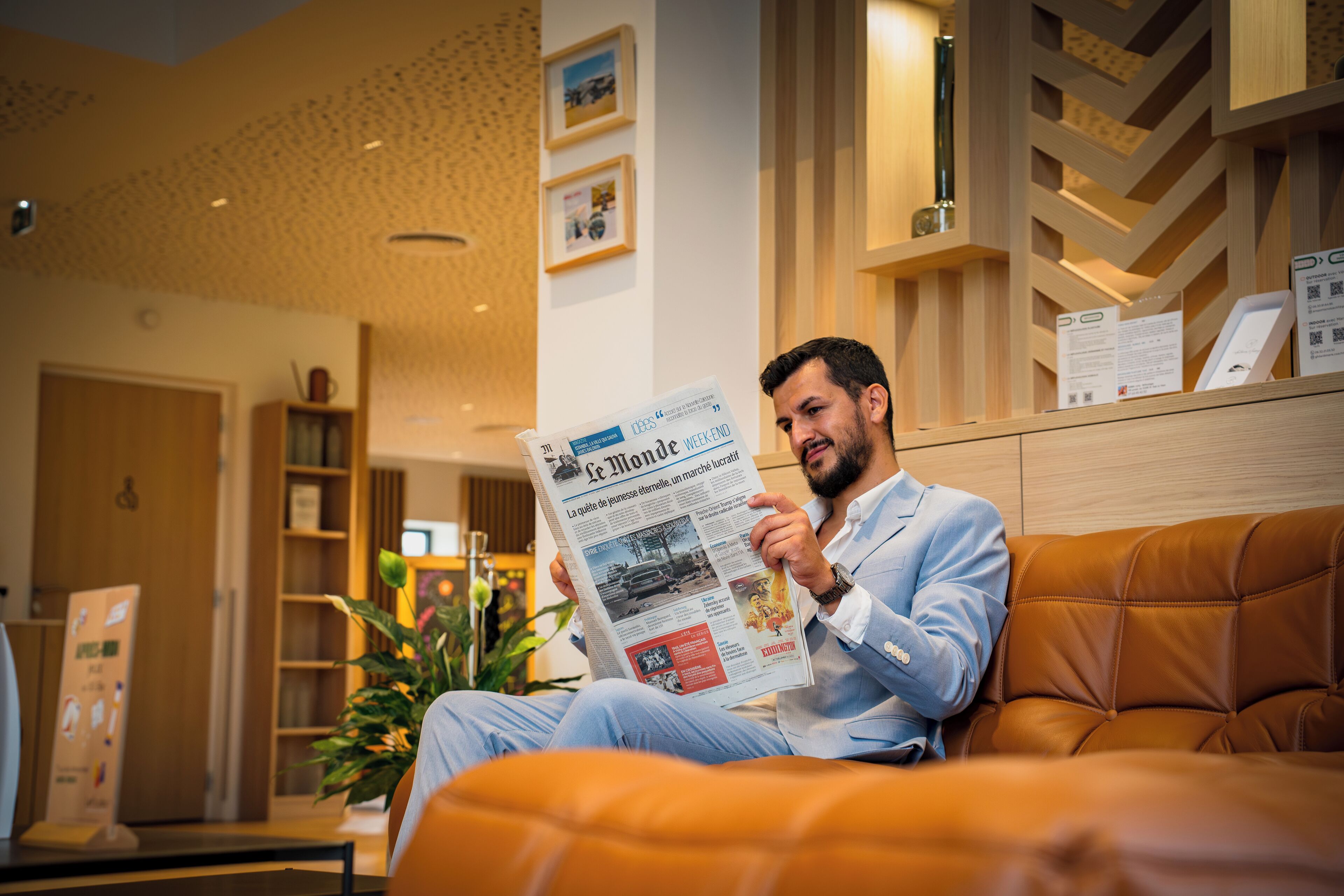 A man reading a newspaper in a modern hotel lobby with a wooden reception desk and a potted plant.