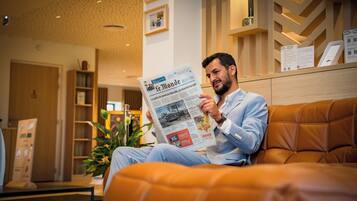A man reading a newspaper in a modern hotel lobby with a wooden reception desk and a potted plant.