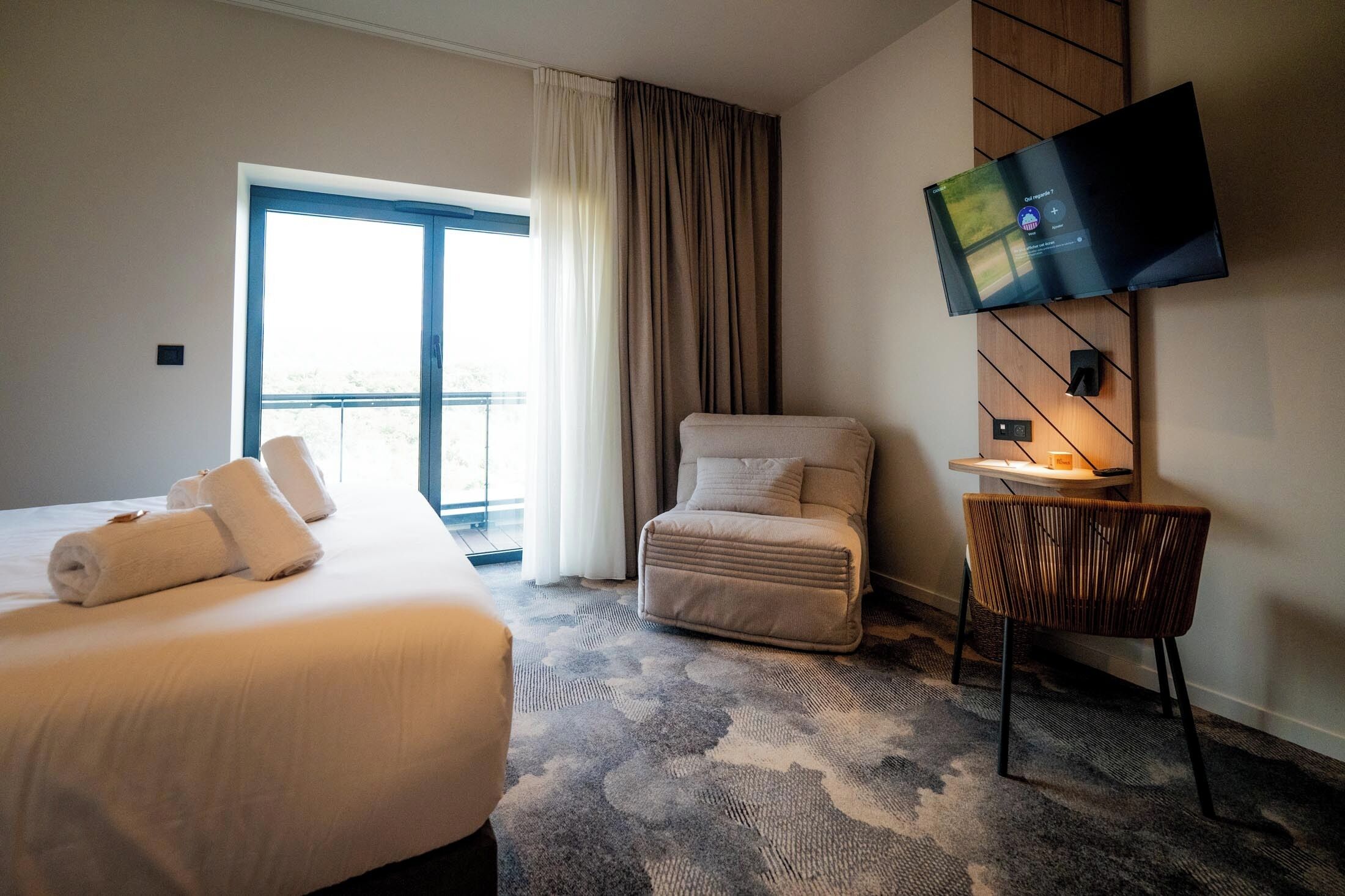 A hotel room with a bed, a chair, a TV mounted on the wall, and a view of the outdoors.
