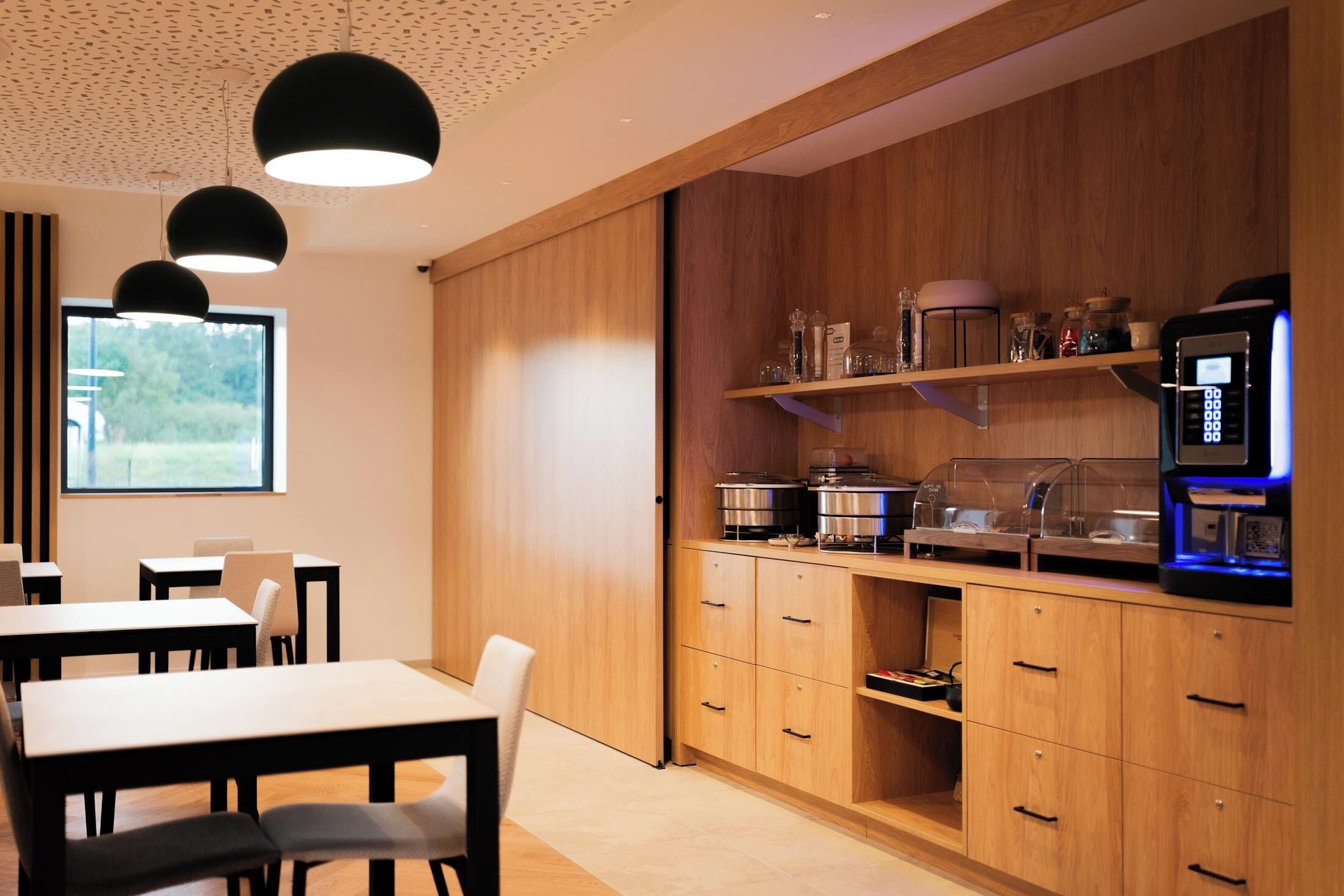 A modern kitchen with wooden cabinets, a coffee machine, and a window with a view of greenery.
