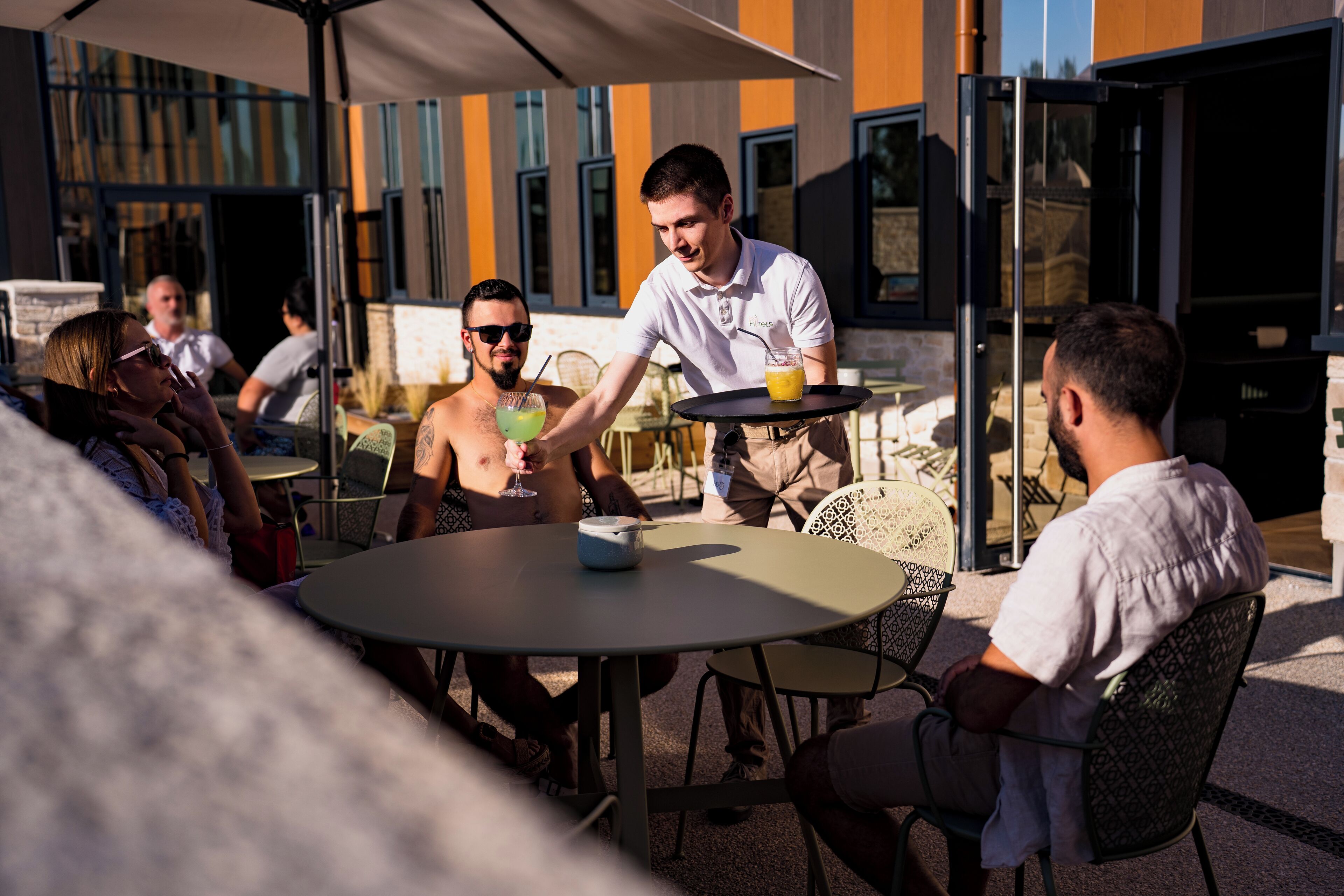 A group of people seated at an outdoor table with a waiter serving drinks.