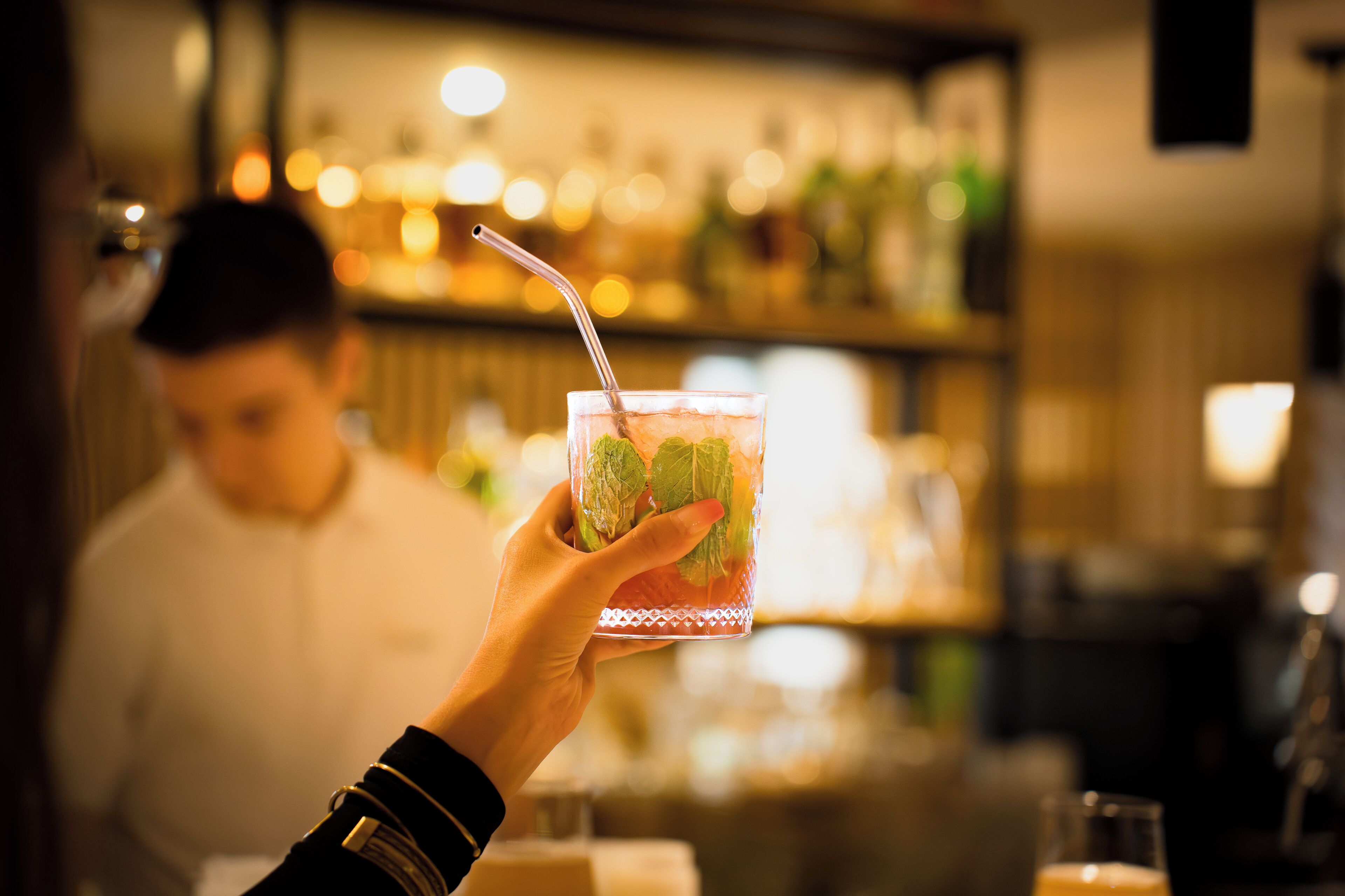 A hand holding a glass with a straw, containing a beverage with mint leaves.
