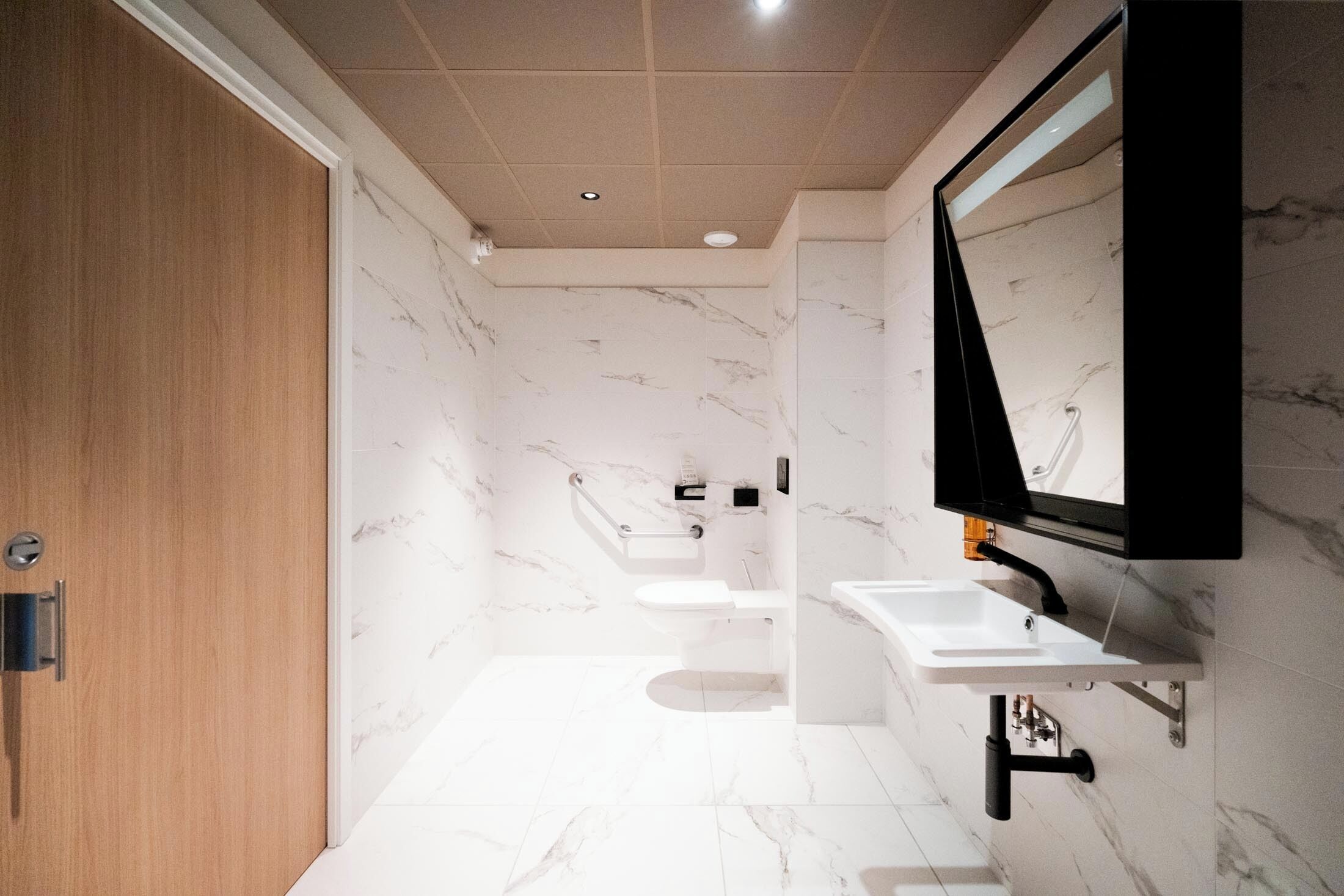 A modern bathroom with marble walls, a wooden door, a large mirror, and a white sink.