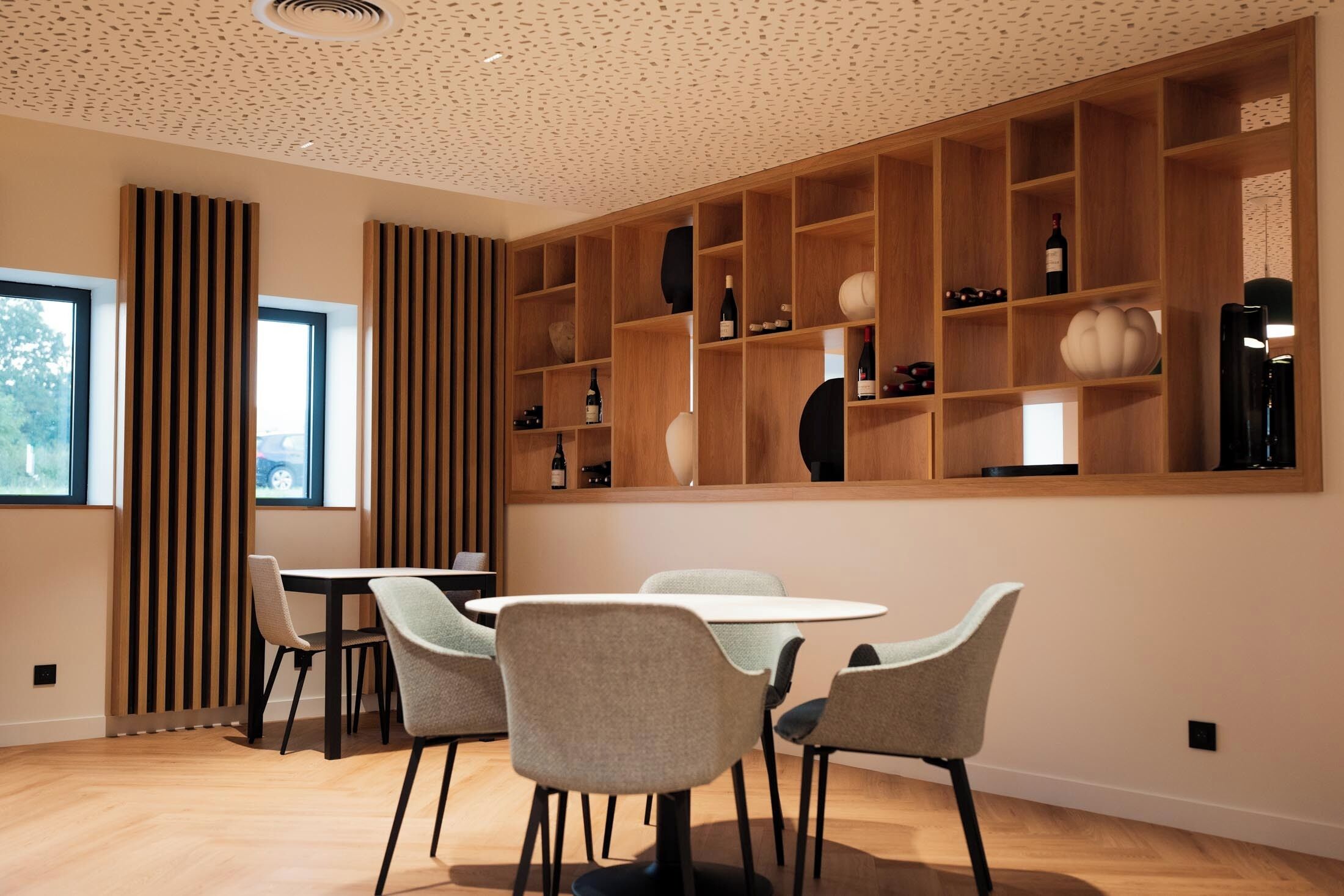 Un espace repas moderne avec une table ronde et quatre chaises, des étagères en bois où sont disposés divers objets, et une fenêtre équipée de stores verticaux.