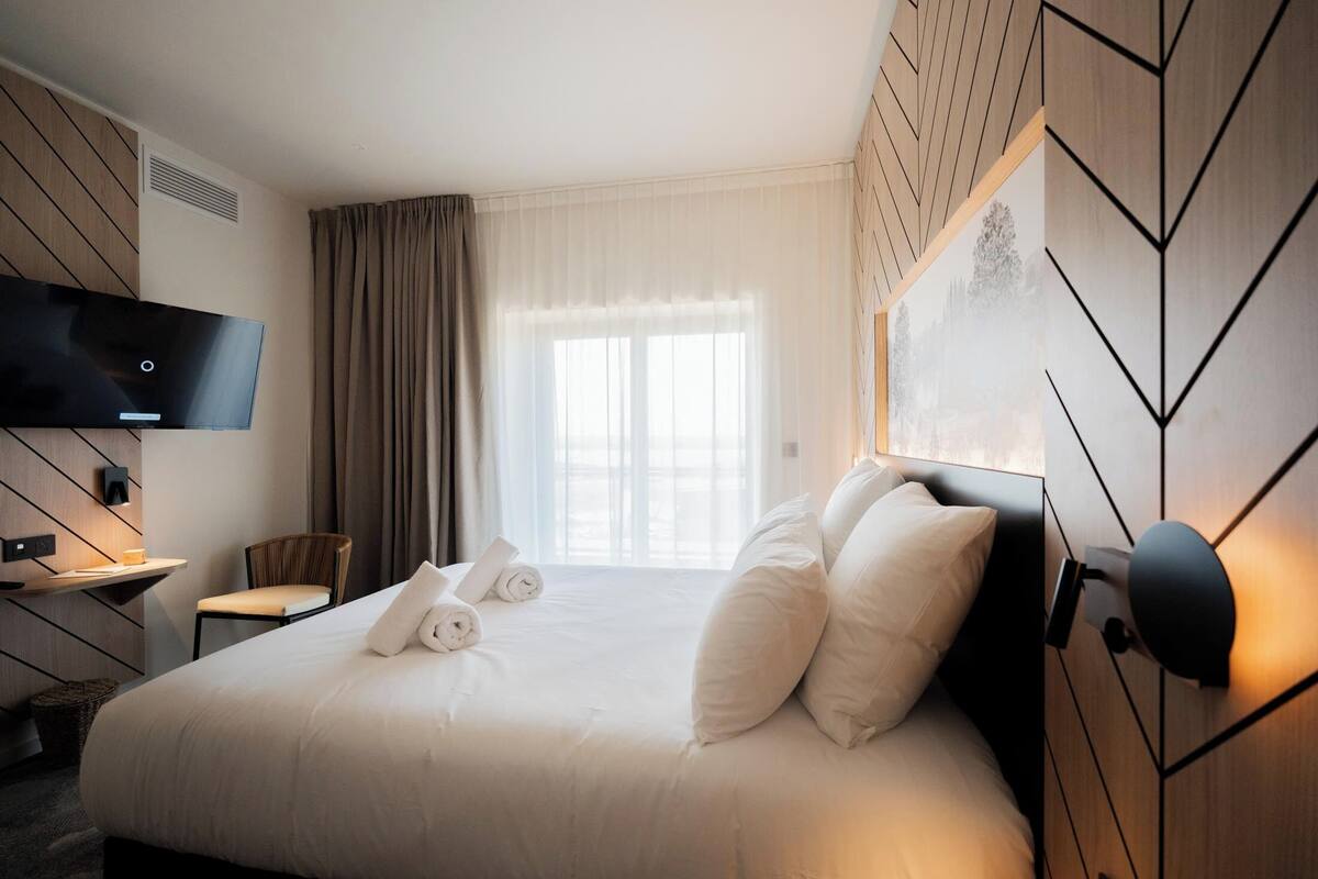 A hotel room with a large bed, a flat-screen TV mounted on the wall, a chair, a desk with a lamp, and a window with curtains.