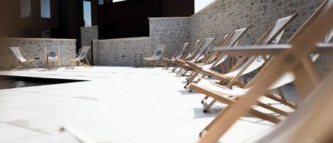 Zona de piscina con tumbonas dispuestas en filas, un muro de piedra y un edificio moderno al fondo.