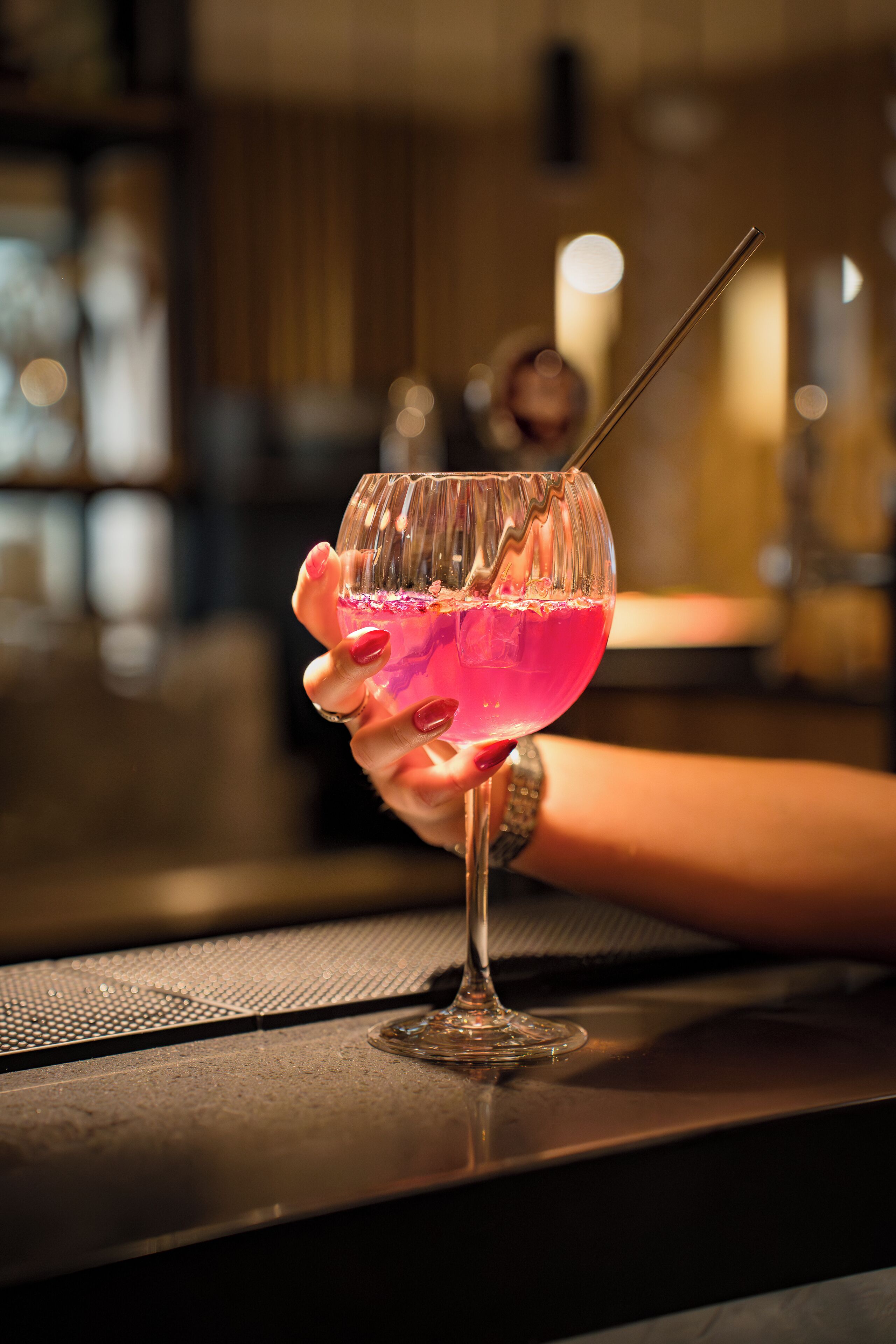 Une main tenant un verre contenant une boisson rose, avec une paille, posé sur le comptoir d’un bar.