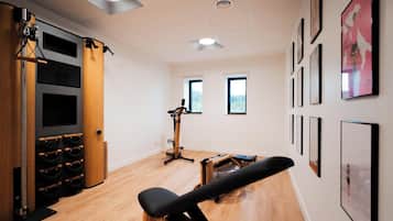 A room with a wooden floor, a black exercise bench, a wall-mounted rack with black and wooden elements, and several framed pictures on the wall.
