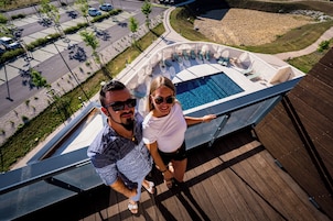 Un hombre y una mujer están de pie en una terraza de madera, con vista a una piscina y sillas de descanso.