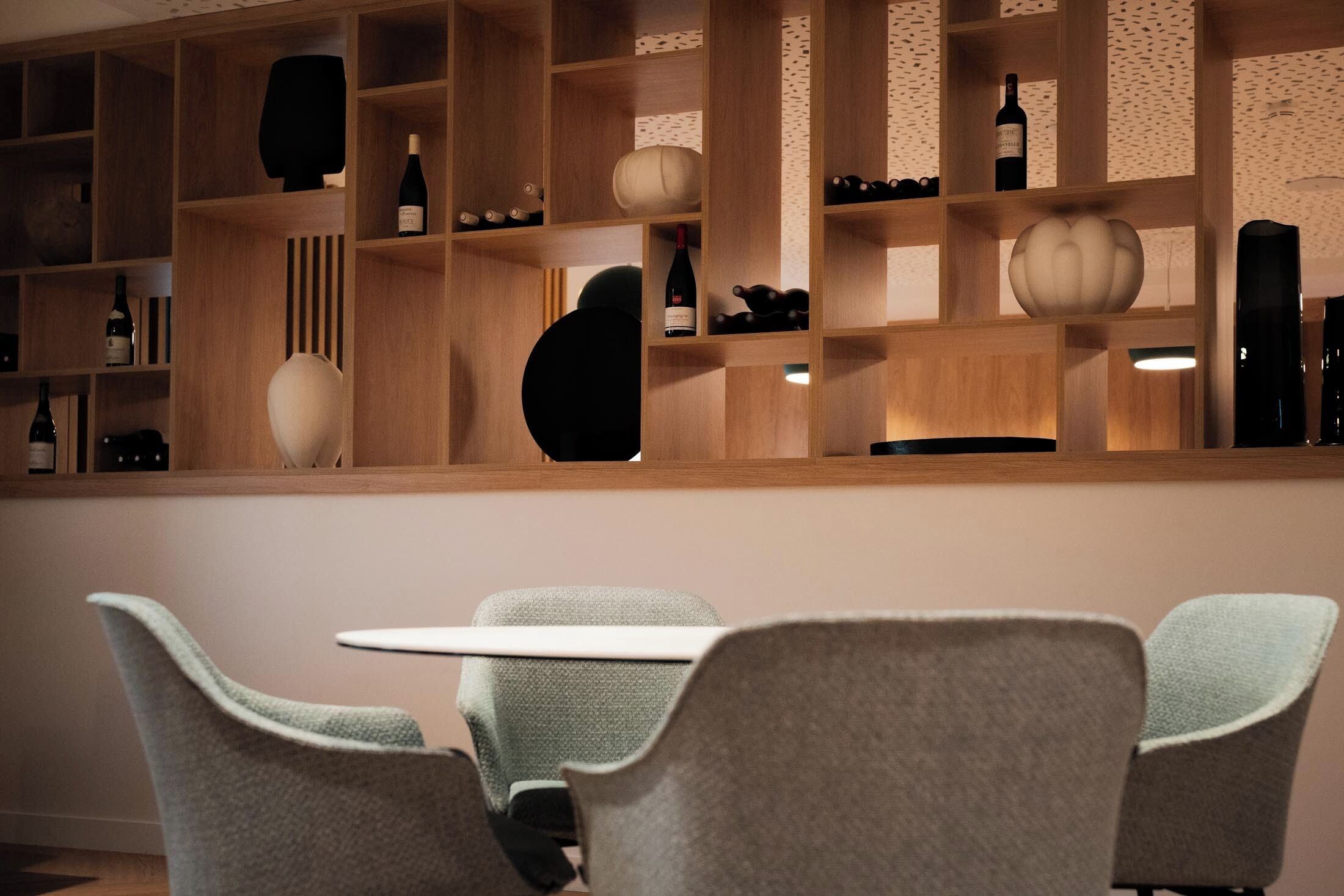 Un interior moderno con una estantería de madera que exhibe diversos objetos, incluyendo botellas y adornos. En primer plano, una mesa redonda con dos sillas.