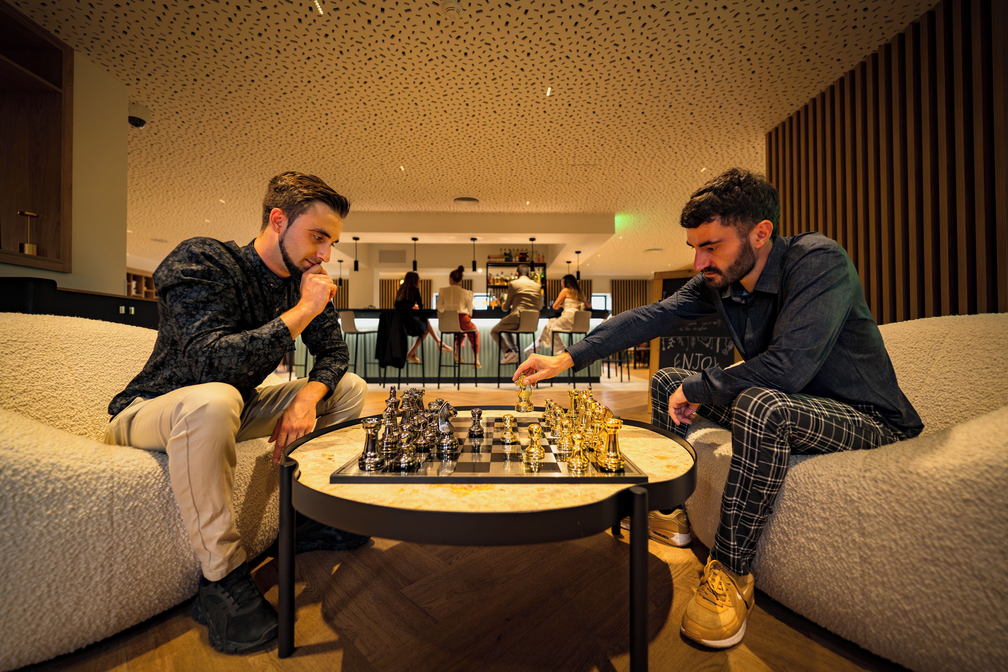 Two men playing chess in a modern lounge area with plush seating and a wooden floor.