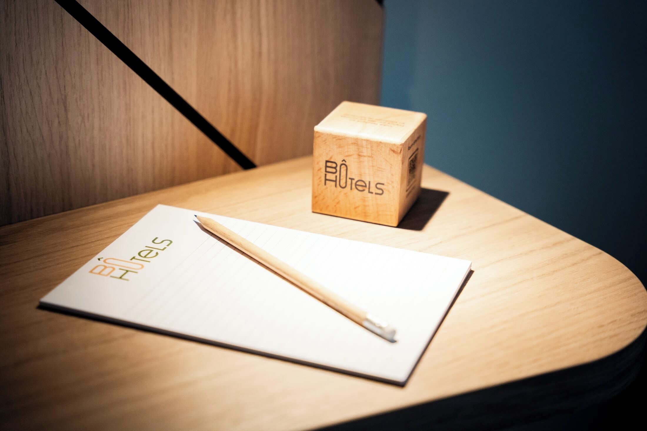 A wooden block with "BóHéis" logo, a white notepad, and a wooden pencil on a wooden surface.