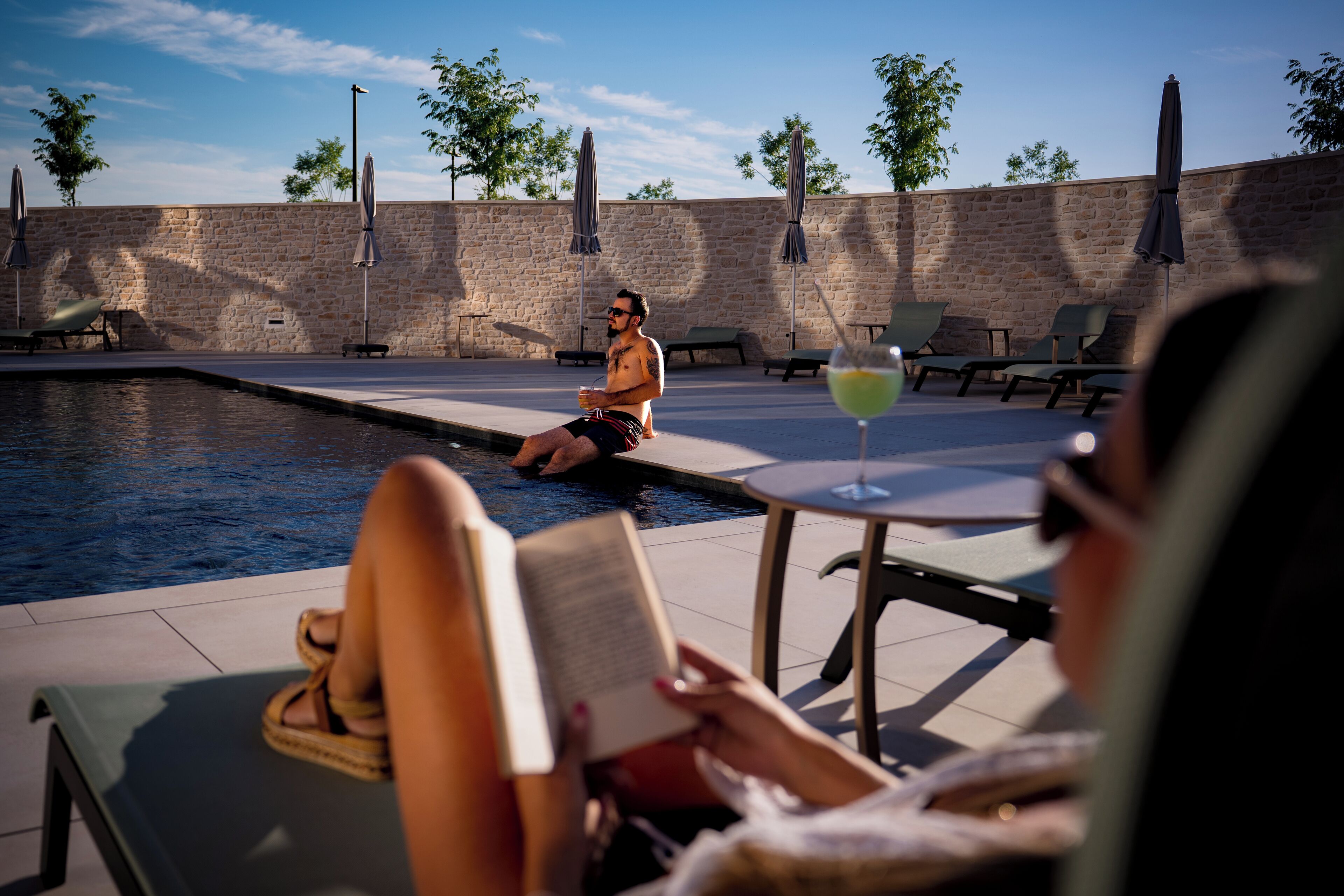Área de piscina com espreguiçadeiras e guarda-sóis, uma parede de pedra e céu claro.