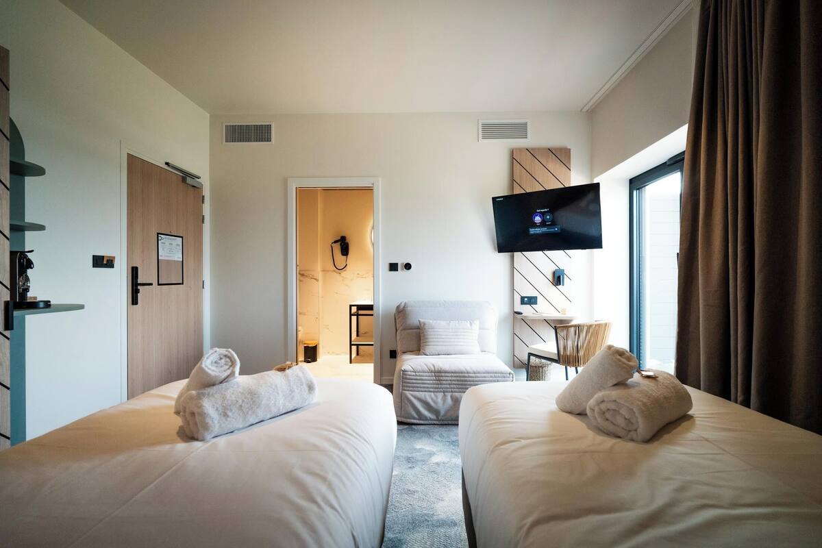 A hotel room with two beds, a TV mounted on the wall, and a view of the outside through the window.