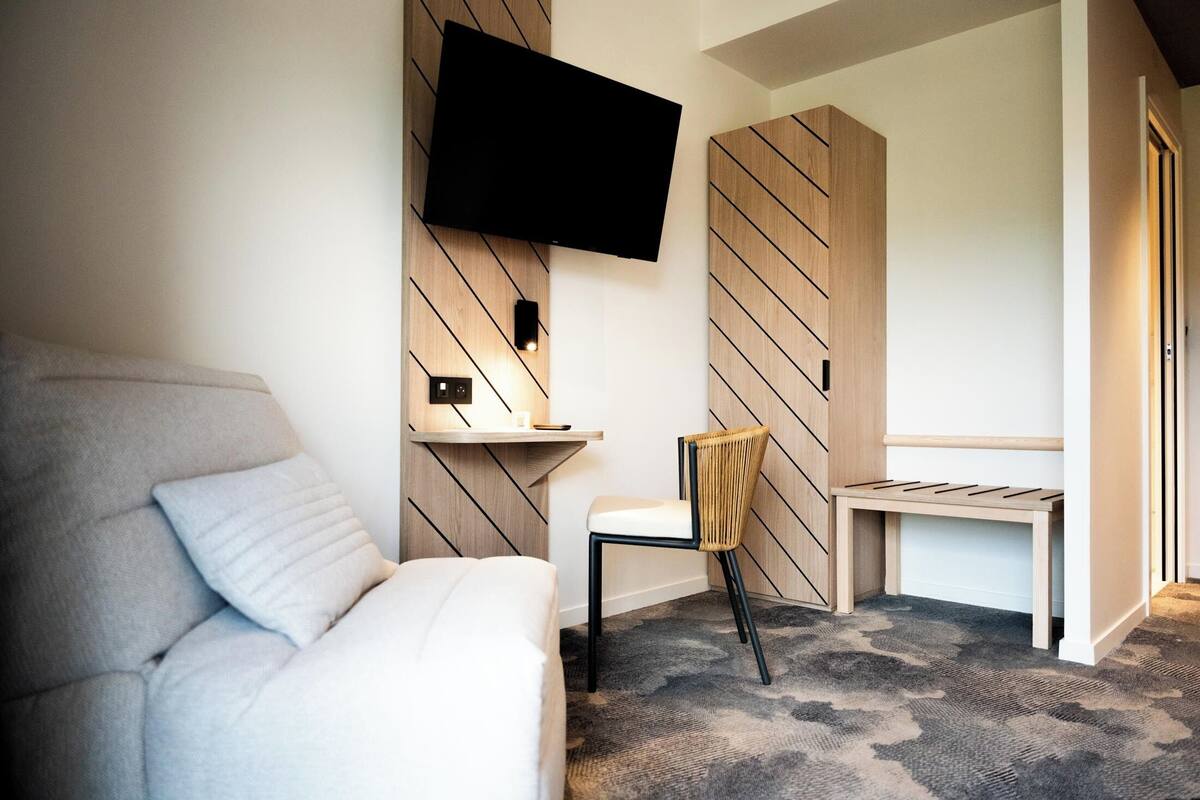 A modern hotel room with a flat-screen TV mounted on the wall, a sofa, a chair, a small table, and a patterned carpet.