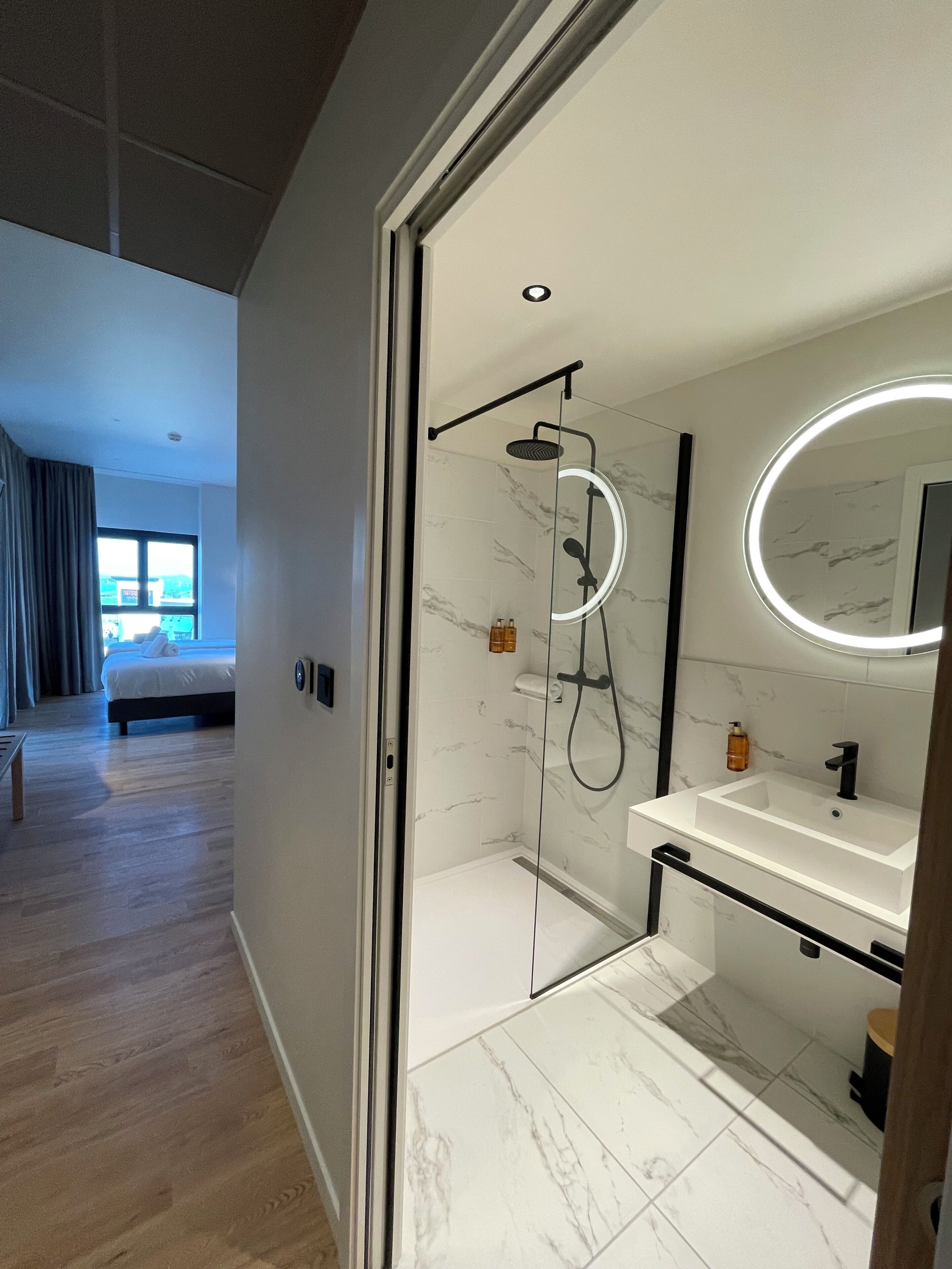 Een moderne badkamer met een douchecabine van glas, een witte wastafel en een grote spiegel met ronde verlichting.