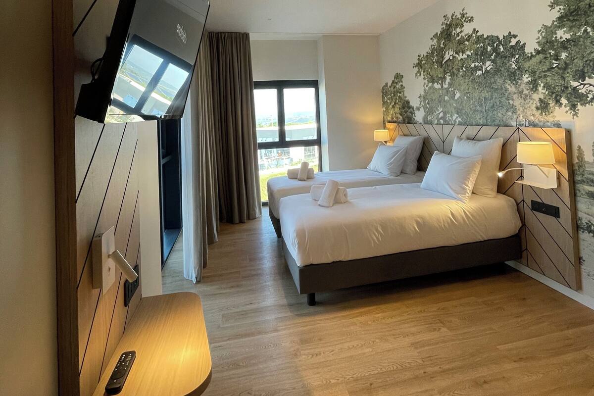 A hotel room with a bed, a TV mounted on the wall, a desk with a remote control, and a window with curtains.