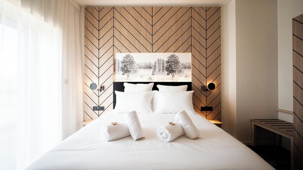 Een net opgemaakt bed met witte lakens en opgerolde handdoeken, een groot abstract schilderij aan de muur, en een houten hoofdbord met een zigzaggend patroon.